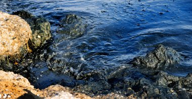Kaya kıyısı, petrol sızıntısı gökkuşağı parıltısıyla kaplı karanlık dalgalarla vuruyor, çevresel felaketin sembolü ve petrol kirliliği sonrasında deniz kirliliğinin yol açtığı deniz yaşamına tehdit oluşturuyor.