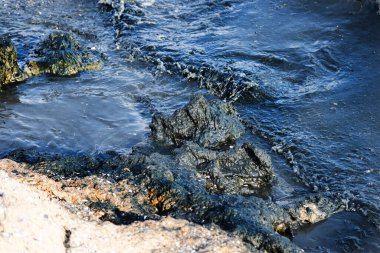 Kaya kıyısı, petrol sızıntısı gökkuşağı parıltısıyla kaplı karanlık dalgalarla vuruyor, çevresel felaketin sembolü ve petrol kirliliği sonrasında deniz kirliliğinin yol açtığı deniz yaşamına tehdit oluşturuyor.