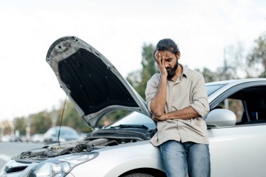 Orta Doğulu bir adam kaputu açık bir şekilde arabasının yanındaki otoparkta duruyor. Parmaklarını saçında gezdirip durumu değerlendirirken hayal kırıklığı belirtileri gösteriyor..