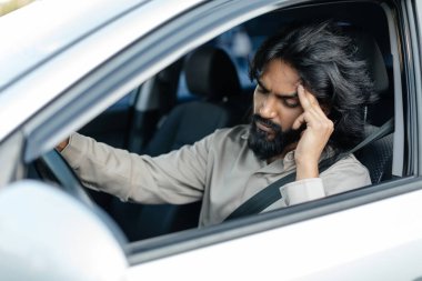 Uzun saçlı ve sakallı bir adam park halindeki bir arabada oturuyor. Stresli ve yorgun görünüyor. Tek eliyle başını tutuyor. Şehir arkaplanı yoğun bir gün olduğunu gösteriyor..