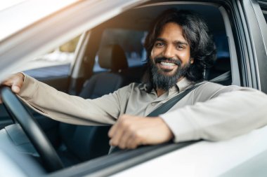 Uzun saçlı ve sakallı genç bir adam kameraya gülümserken arabasında rahat bir şekilde oturmaktan hoşlanıyor. Pencereden güneş ışığı süzgeçleri.