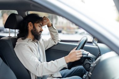 Uzun saçlı ve sakallı bir adam bir arabanın direksiyonunun başında sıkıntılı görünüyor. Şehir trafiğinde yol alırken direksiyonu sıkıca tutuyor..