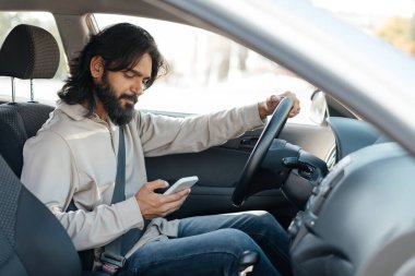  Ortadoğulu bir adam bir arabanın şoför koltuğunda oturur ve kemerlerini bağlayıp akıllı telefonuna odaklanır. Ayar, güneşli ve güneşli bir günü gösteriyor. Bu da gündelik bir sürüş olduğunu gösteriyor..