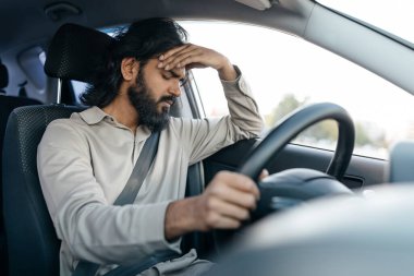 Orta Doğulu bir adam endişeli bir ifadeyle direksiyonun başında oturur, direksiyonu sıkıca tutar. Düşüncelere dalmış gibi görünüyor, şehirde araba sürerken stresi yansıtıyor..