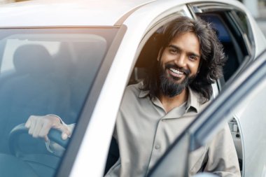 Neşeli bir adam açık renkli bir gömlek giyerek arabasının camından dışarı sarkmış. Güneş ışığı yüzünü aydınlatır, günün tadını çıkarırken, kaygısız bir ruhu somutlaştırır..