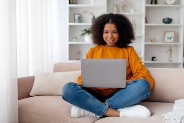 Minimalist oturma odasında bilgisayarda yazı yazan neşeli, güzel, Afro-Amerikan bir kadın. Çalışma, serbest çalışma, iş ve iş uzaktan evde, cihaz ve yeni normal. 