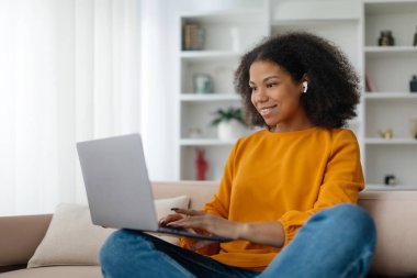 Afro-Amerikalı serbest çalışan, dizüstü bilgisayar kullanan ve bilgisayarında çalışan kablosuz kulaklıklar takan evdeki oturma odasındaki kanepede oturan bir kadın. Serbest Yaşam Tarzı Konsepti. Yüksek kalite fotoğraf