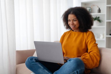 Evdeki kanepede oturan, dizüstü bilgisayar kullanan, internetten video izleyen, arkadaşlarıyla sohbet eden, blog okuyan, fotokopi çeken Afro-Amerikan bir bayan. Covid-19 salgını sırasında yaşam tarzı. 