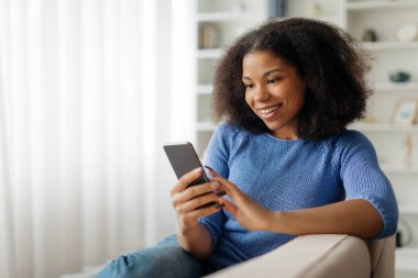 Evde cep telefonu kullanan mutlu siyahi kadının portresi, akıllı telefondan alışveriş yapan veya arkadaşlarıyla mesajlaşan gülümseyen Afro-Amerikan bayan, koltukta dinlenirken yeni uygulamalara göz gezdirirken....