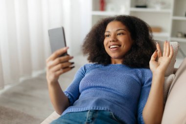 Çevrimiçi İletişim. Mutlu Siyahi Kadın Evdeki Akıllı Telefon vasıtasıyla Arıyor, Kanepede oturan ve el sallayan Genç Afrikalı Amerikalı Kadın, Cep Telefonu Kamerası,. 