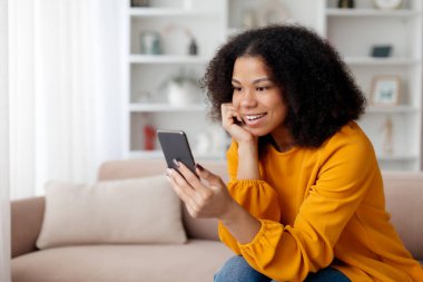 Neşeli siyahi kadın evde koltukta dinlenirken cep telefonu kullanıyor arkadaşlarıyla mesajlaşıyor ya da sosyal medyayı tarıyor, boşluğu kopyalıyor. Yüksek kalite fotoğraf