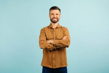 A cheerful caucasian middle aged man stands with arms confidently crossed, wearing a casual brown shirt, against a solid blue background. High quality photo clipart