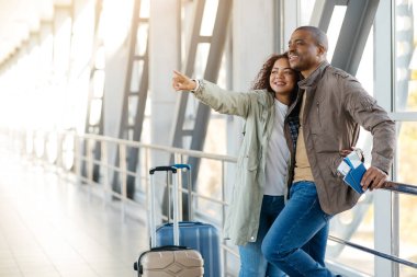 En iyi havayolları konsepti. Havaalanındaki mutlu Afrikalı Amerikalı çift fotokopi uzayını işaret ediyor, Afrikalı Amerikalı eşler bavullarıyla terminalde yürüyor, balayı tatiline hazır..