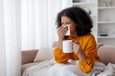 Genç Afro-Amerikan kadın soğuk algınlığı geçiriyor, burnunu silip elinde çay tutuyor, hasta Afro-Amerikan kadın evde oturuyor, grip belirtileri gösteriyor, fotokopi çekiyor. Yüksek kalite fotoğraf