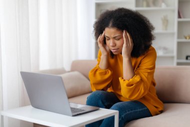 Hastalığın ilk belirtileri, migren, aşırı çalışma. Üzgün, genç, Afro-Amerikalı bir kadın dizüstü bilgisayarla koltukta oturuyor. Alnına baskı yapıyor. Oturma odasında baş ağrısı çekiyor. Yüksek kalite fotoğraf