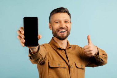 Pozitif Avrupalı orta yaşlı adam elinde boş ekranla akıllı telefonu işaret ediyor ve gülümsüyor, mavi stüdyo arka planında izole edilmiş, yeni online teklif sunuyor, maket. Yüksek kalite fotoğraf