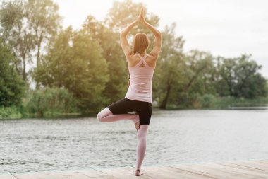Huzurlu bir ortamda, bir kadın bir bacağının üzerinde dengede duruyor. Yoga pozu veriyor. Huzurlu sular ve berrak bir gökyüzünün altında canlı yeşilliklerle çevrili..