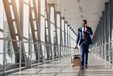 Takım elbiseli bir adam gösterişli bir havaalanı terminalinde güvenle yürüyor, tekerlekli bavulu çekiyor ve öğleden sonra telefonuyla meşgul oluyor..