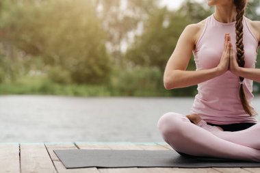 A woman sits cross-legged on a yoga mat by the water, hands in a prayer position, enjoying a peaceful moment of meditation during sunset, surrounded by nature. clipart