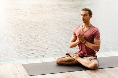 A man with a focused expression is meditating in a seated position on a yoga mat beside a tranquil body of water during sunset. clipart