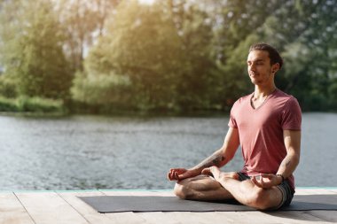 Huzurlu bir gölün yakınındaki yoga minderinde oturan bir adam, parlak güneş ışığı altında yemyeşil bir yeşillikle çevrelenmiş bir şekilde meditasyonun tadını çıkarıyor..