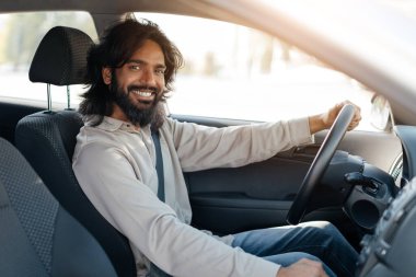 Genç bir adam güneşli bir günde mutlu bir şekilde arabasını sürüyor. Neşe dolu bir ifade sergiliyor, direksiyonu tutarken sürücü koltuğuna doğru gevşiyor..