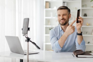 Sıradan tişörtlü bir adam internette akıllı telefonunu gösterip gülümsüyor. Elinde bir dizüstü bilgisayar ve mikrofonla çalışma masasında oturuyor..