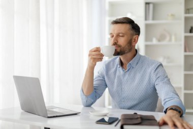 Sakallı bir adam masada oturup bir fincan kahvenin tadını çıkarıyor. Dizüstü bilgisayarına odaklanmış, iyi aydınlatılmış modern ofis ortamında çalışmaya dalmış..