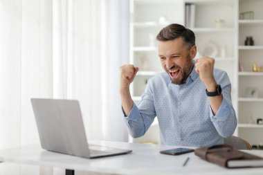 Bir adam iyi ışıklandırılmış, çağdaş bir ofis ortamında dizüstü bilgisayarı üzerinde çalışırken bir hedefe ulaştıktan sonra sevinç ve heyecan ifade eder..