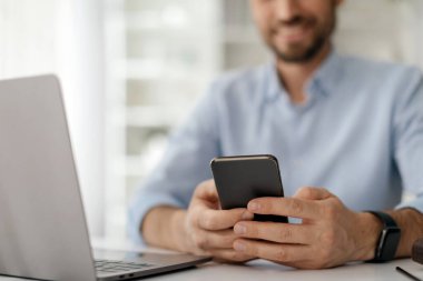 Bir adam akıllı telefonuyla ilgilenirken aynı zamanda iyi ışıklandırılmış bir ofis ortamında dizüstü bilgisayarla çalışıyor..