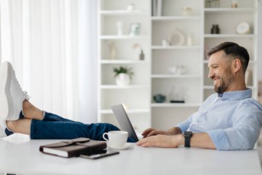 Ayakları havada bir masada rahat bir şekilde oturan bir adam şık bir ev ofisinde dizüstü bilgisayarla çalışıyor. Güneş ışığı perdelerin arasından süzülüp rahat bir ortam yaratır..