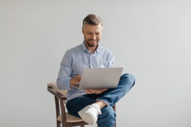 Sakallı bir adam dizüstü bilgisayarıyla çalışmaktan hoşlanır, açık mavi gömlek ve günlük kot pantolon giyer. İyi ışıklandırılmış, minimalist bir odada rahat rahat oturuyor..