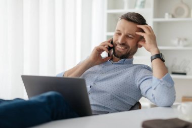 Bir adam evinde dinleniyor, bilgisayarını aktif olarak kullanırken telefonuyla sohbet ediyor. Sabah güneşi odayı dolduruyor..