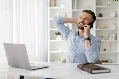Bir adam rahat bir şekilde masada oturup, rahat bir ifadeyle telefonda konuşuyor. Önüne dizüstü bilgisayarı ve bir defter konur. Günışığı modern odayı doldurur..