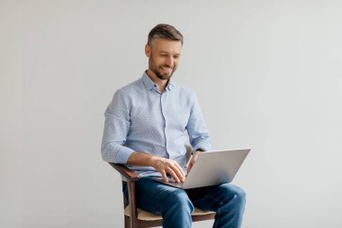 Bir adam, parlak ve havadar bir çalışma alanında ahşap bir sandalyede otururken dizüstü bilgisayarına derinden odaklanır. Tarafsız arka plan konsantrasyonunu arttırıyor..