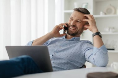 Adam, rahat ve neşeli, masasında bir telefon görüşmesi yapıyor, doğal ışık ve çağdaş dekor ile çevrili, verimli bir çalışma ortamı gösteriyor.