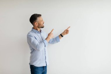 Açık mavi gömlekli ve kot pantolonlu bir adam iki eliyle boş duvara doğru işaret ediyor, iyi ışıklandırılmış bir ev ortamında coşkulu görünüyor..