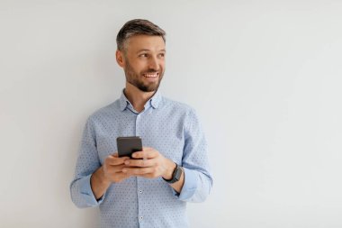 Sakallı neşeli bir adam düz bir duvara yaslanmış, akıllı telefonuyla meşgul, açık mavi bir gömlek giymiş ve memnun görünüyor..