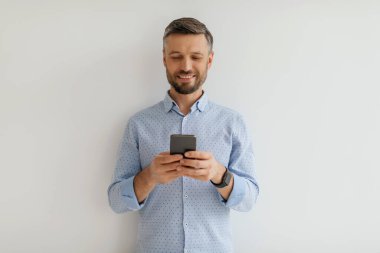 Bir adam akıllı telefonuyla uğraşırken ışık duvarına yaslanmış gülümsüyor. Parlak atmosfer neşeli bir davranışı yansıtıyor, bir anlık mutluluğu işaret ediyor..