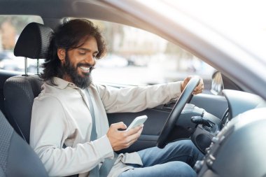 Bir adam araba sürerken, akıllı telefonuna bakarken gülümseyerek bir anlık rahatlıktan zevk alır. Neşeli atmosfer parlak gün ışığı ve kentsel çevre ile yükseldi..
