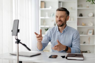 Açık mavi gömlekli bir adam evinde sanal bir toplantı düzenliyor. Akıllı telefon ve dizüstü bilgisayarla iletişim kurarken sürekli gülümsüyor ve hareketler yapıyor..