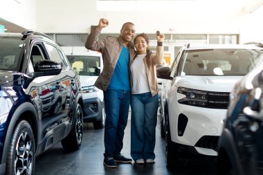 Genç bir Afrikalı Amerikalı çift bir galeride araba alımlarını tamamladıktan sonra neşeyle gülümsüyor. Onlar galeride çeşitli araçlarla çevrilidir..