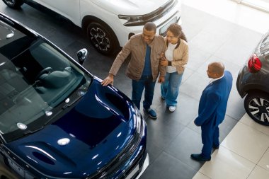 Genç bir Afrikalı Amerikalı çift galeride bir arabayı incelerken satış seçeneklerini değerlendiren bir satıcıyla ayrıntıları tartışıyor..