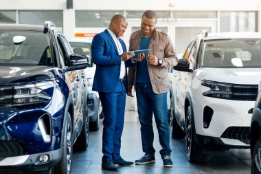 Genç Afrikalı Amerikalı bir adam, çeşitli otomobillerle dolu modern bir galeride bir araba satıcısıyla araç özelliklerini tartışıyor. Satın alma veya kiralama seçeneklerini incelerler.