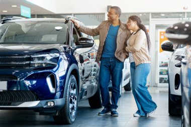 Genç bir Afrikalı Amerikalı çift araba galerisinde potansiyel araba alımları hakkında konuşuyorlar. Gülümserken bir aracı incelerler..