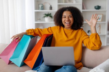 Kıvırcık saçlı genç bir kadın evde rahatça oturuyor, gülümsüyor ve online alışveriş yaparken el sallıyor. Renkli alışveriş torbaları onun yanına yerleştiriliyor, boş zamanlarını sergiliyor..