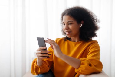 Akıllı telefonlu ve kablosuz kulaklıklı genç siyahi kadın evde dinleniyor, neşeli Afro-Amerikan kadın müzik dinliyor ve oturma odasındaki kanepede dinlenirken cep telefonuyla internette geziniyor.