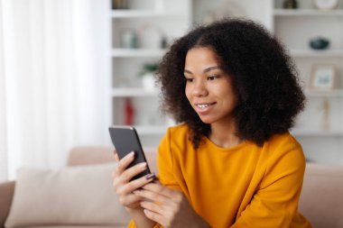 Genç Afro-Amerikalı bir kadın evinde rahat bir şekilde otururken akıllı telefon kullanıyor, cep telefonuyla konuşurken gülümsüyor, internetten alışveriş yapıyor ya da sohbet ediyor.