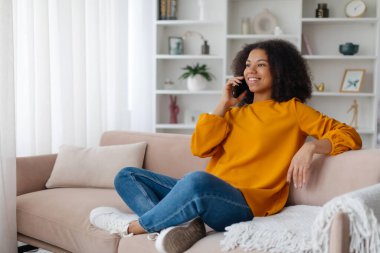 Genç Afrikalı Amerikalı bir kadın evinde cep telefonuyla konuşuyor, kanepede oturuyor, arkadaşlarıyla telefonda konuşuyor, boş zamanlarında neşe saçıyor.