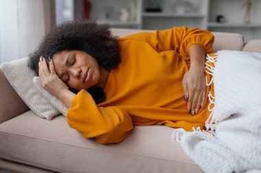 Hasta siyah kadın kanepede yatıyor, kendini kötü hissediyor, hasta Afrikalı Amerikalı kadın adet sancısı çekiyor, üzgün kadın karın ağrısı ve şiddetli baş ağrısı çekiyor.